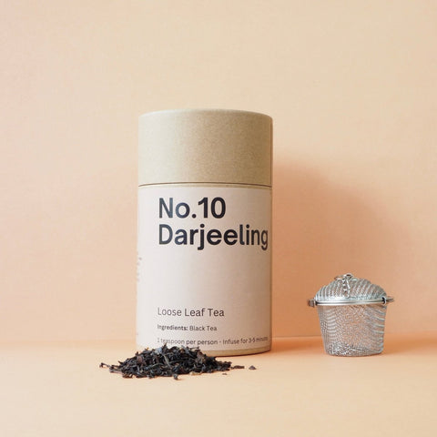 A cylindrical brown packaging labeled 'No.10 Darjeeling Loose Leaf Tea', with a small pile of loose leaf tea to the left and a metal tea strainer beside it, all against a pink background.