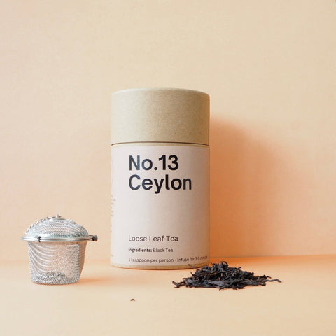 A cylindrical container labeled 'No.13 Ceylon' with loose leaf tea scattered in front and a metal tea strainer to the side, all against an orange background.