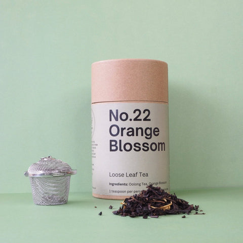 A cylindrical brown paper container labeled 'No.22 Orange Blossom Loose Leaf Tea', with a silver tea strainer and loose tea leaves scattered in front, all against a light green background.