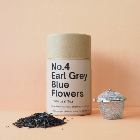 A cylindrical container labeled 'No.4 Earl Grey Blue Flowers Loose Leaf Tea' is displayed alongside a strainer and a scattering of dried tea leaves on a peach background.