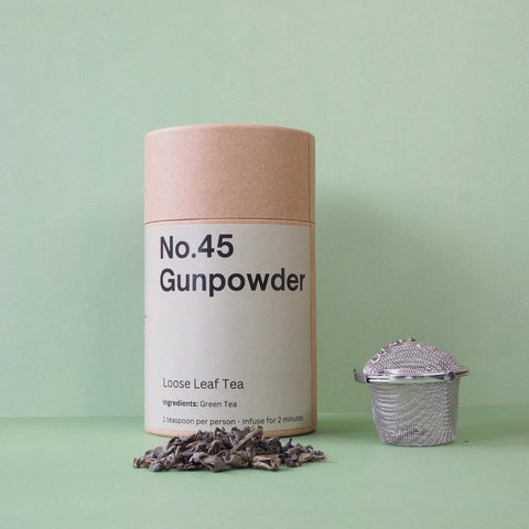 A cylindrical paper container labeled 'No.45 Gunpowder Loose Leaf Tea' on a green background, alongside a metal tea strainer and scattered dry tea leaves.