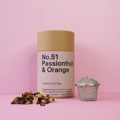 A cylindrical paper container labeled 'No.51 Passionfruit & Orange Loose Fruit Tea' accompanied by a metal tea strainer and a scattering of dried fruit pieces on a pink background.