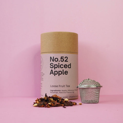 A cylindrical cardboard container labeled 'No.52 Spiced Apple' next to a metallic tea strainer and scattered dried apple pieces on a pink background.