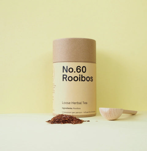 A cylindrical paper container labeled 'No.60 Rooibos' with loose herbal tea leaves scattered in front and a small wooden spoon to the right.