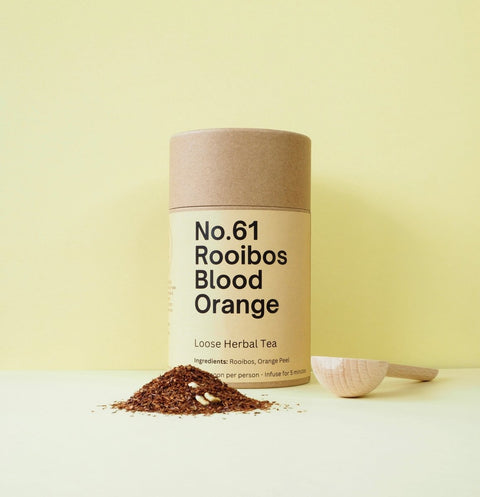 A cylindrical container labeled 'No.61 Rooibos Blood Orange' with loose herbal tea leaves scattered in front and a scoop to the side on a yellow background.