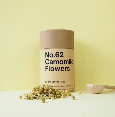 A cylindrical container labeled 'No.62 Camomile Flowers' with loose herbal tea leaves scattered in front, on a yellow background.