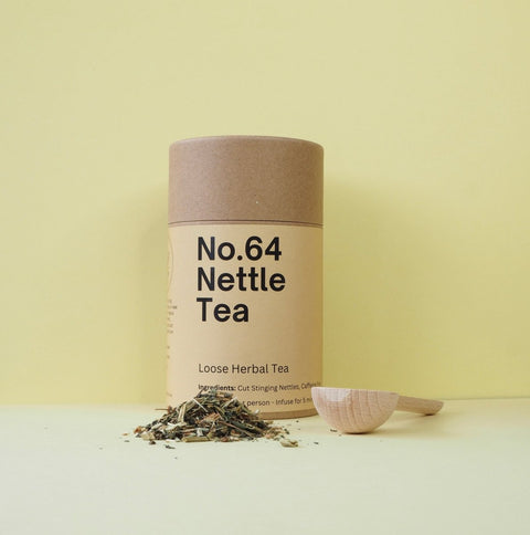 A cylindrical paper container labeled 'No.64 Nettle Tea' next to a wooden spoon with loose herbal tea leaves scattered around.