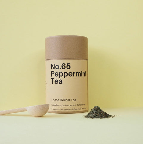 A cylindrical paper container labeled 'No.65 Peppermint Tea' with a wooden spoon beside it, and loose peppermint tea leaves scattered in front, all against a yellow background.