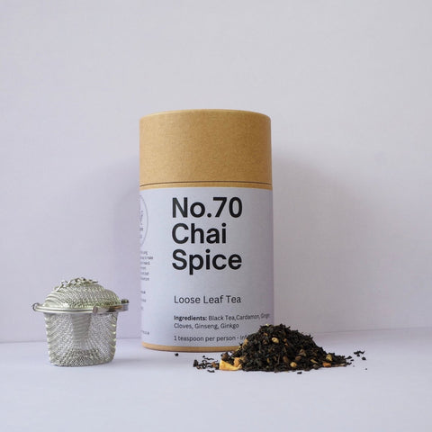 A cylindrical brown cardboard packaging labeled 'No.70 Chai Spice Loose Leaf Tea' with a silver tea ball and a pile of loose tea leaves in the foreground.