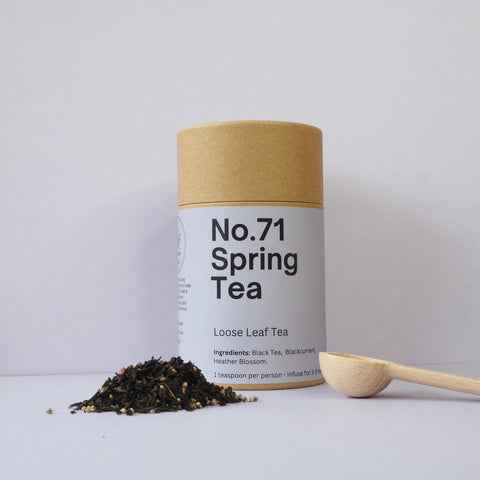 A cylindrical brown container labeled 'No.71 Spring Tea' next to a wooden spoon with loose leaf tea scattered in front.