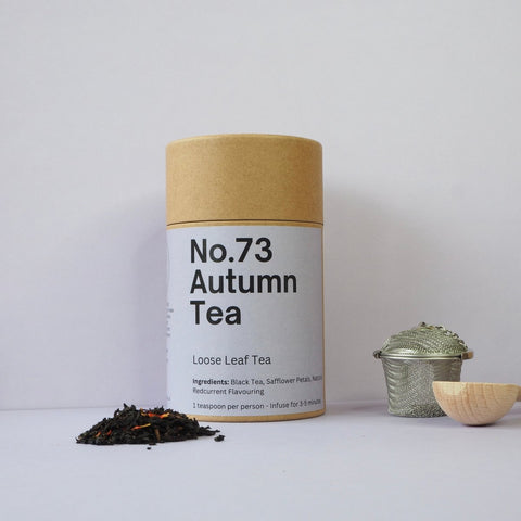 A cylindrical paper container labeled 'No.73 Autumn Tea' next to a small metal strainer and a pile of loose leaf tea on a white background.