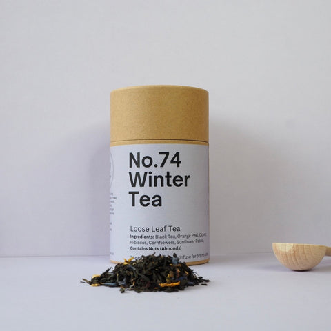 A cylindrical brown paper tube packaging labeled 'No.74 Winter Tea', with loose leaf tea visible in front and a wooden spoon to the right.