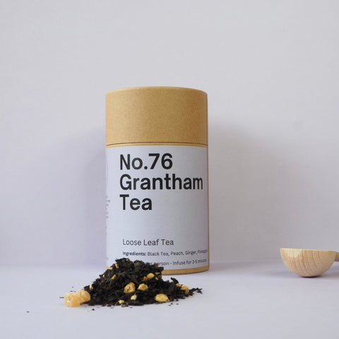 A cylindrical container labeled 'No.76 Grantham Tea' with a beige lid, next to a scattered pile of loose leaf tea with visible ginger pieces.