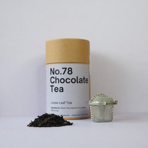 A cylindrical brown container labeled 'No.78 Chocolate Tea' next to a glass tea ball and a pile of loose leaf tea on a white background.