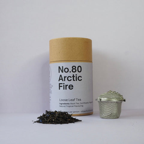 A cylindrical container labeled 'No.80 Arctic Fire' next to a pile of loose leaf tea and a metal tea strainer.