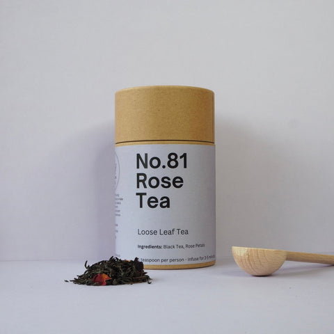 A cylindrical paper container labeled 'No.81 Rose Tea' with a wooden spoon beside it, and loose tea leaves scattered in front.