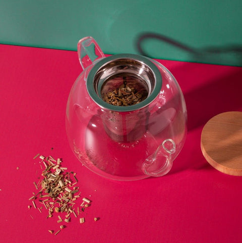 Clear glass teapot with metal infuser on a red surface, surrounded by loose tea leaves.