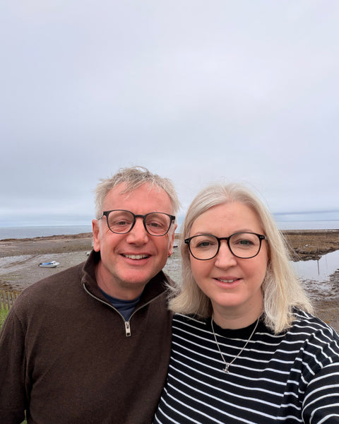 A man and a woman wearing black-rimmed glasses taking a selfie outdoors against an overcast sky and coastal background.