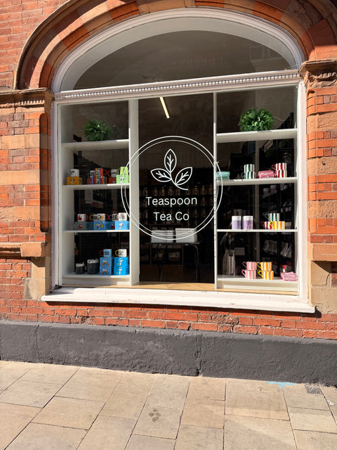 A storefront window of a tea shop with brick walls, displaying shelves of packaged tea products and a large white 'Teaspoon Tea Co' logo on the glass.