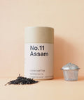 A cylindrical container labeled 'No.11 Assam' next to scattered loose leaf tea and a metal tea strainer on a peach background.