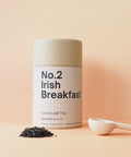 A cylindrical tin container labeled 'No.2 Irish Breakfast' next to a wooden spoon with loose leaf tea scattered in front, all against a pink background.