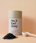 A cylindrical tin container labeled 'No.3 Earl Grey Loose Leaf Tea' with a wooden spoon and a pile of loose tea leaves in front of it, all against a peach background.