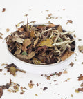 A bowl of Pai Mu Tan white loose leaf tea on a white background, surrounded by scattered dried tea leaves.
