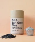 A cylindrical container labeled 'No.4 Earl Grey Blue Flowers Loose Leaf Tea' is displayed alongside a strainer and a scattering of dried tea leaves on a peach background.