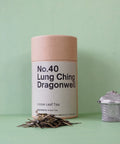 A cylindrical paper container labeled 'No.40 Lung Ching Dragonwell Loose Leaf Tea' positioned next to a metal tea strainer, with loose tea leaves scattered in front.