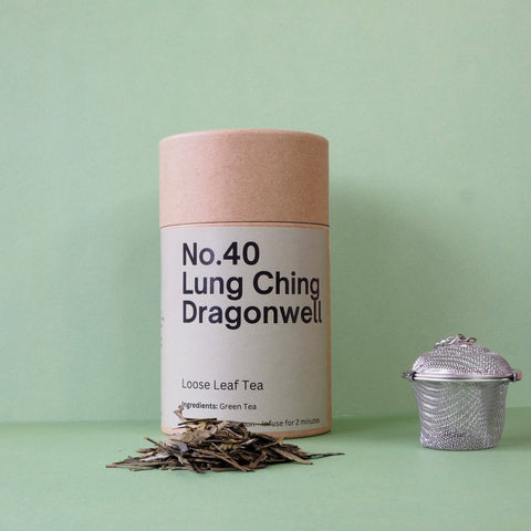 A cylindrical paper container labeled 'No.40 Lung Ching Dragonwell Loose Leaf Tea' positioned next to a metal tea strainer, with loose tea leaves scattered in front.