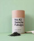 A cylindrical paper container labeled 'No.42 Sencha Fukujyu Loose Leaf Tea' with a wooden spoon and loose tea leaves scattered in front on a green background.