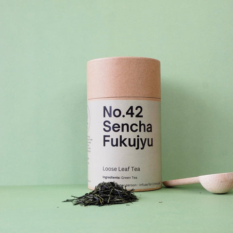 A cylindrical paper container labeled 'No.42 Sencha Fukujyu Loose Leaf Tea' with a wooden spoon and loose tea leaves scattered in front on a green background.