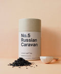 A cylindrical tin container labeled 'No.5 Russian Caravan Loose Leaf Tea' on a pink background, with loose black tea leaves scattered in front 