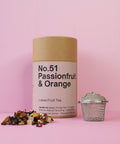 A cylindrical paper container labeled 'No.51 Passionfruit & Orange Loose Fruit Tea' accompanied by a metal tea strainer and a scattering of dried fruit pieces on a pink background.