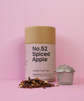 A cylindrical cardboard container labeled 'No.52 Spiced Apple' next to a metallic tea strainer and scattered dried apple pieces on a pink background.
