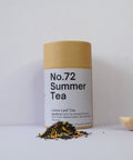 A cylindrical container labeled 'No.72 Summer Tea' with a kraft paper lid, next to a small wooden bowl and loose tea leaves scattered in the foreground.
