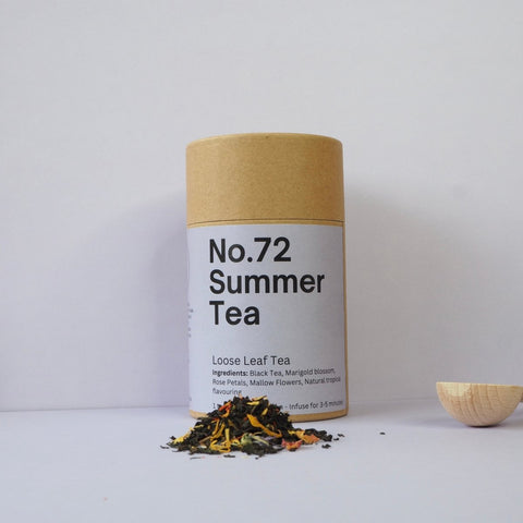 A cylindrical container labeled 'No.72 Summer Tea' with a kraft paper lid, next to a small wooden bowl and loose tea leaves scattered in the foreground.