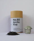 A cylindrical container labeled 'No.80 Arctic Fire' next to a pile of loose leaf tea and a metal tea strainer.
