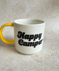 White mug with yellow handle and 'Happy Camper' text on a beige surface