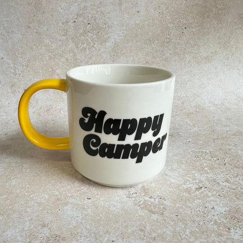 White mug with yellow handle and 'Happy Camper' text on a beige surface
