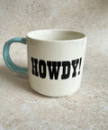 White mug with 'HOWDY!' printed on it, sitting on a textured surface.