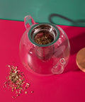 Clear glass teapot with metal infuser on a red surface, surrounded by loose tea leaves.