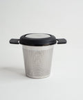 A stainless steel cup infuser for loose leaf tea with a black lid and handle, displayed against a white background.